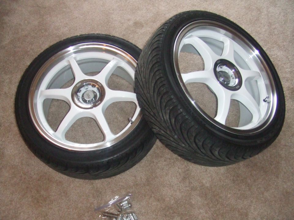Two white 5-spoke wheels with tires on a carpeted surface. A Supermotors.net logo is visible