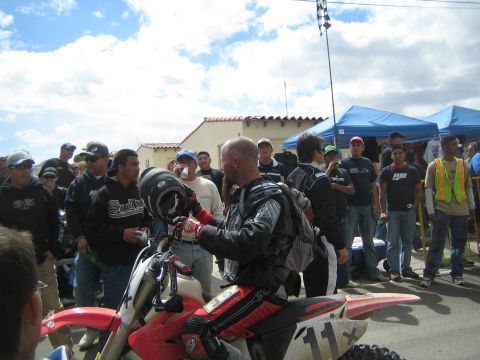1990 Ford Bronco - rider in black leather gear steadies a red dirt bike among a crowd as a checkered flag waves above.