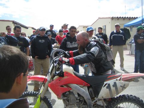 1990 Ford Bronco - bearded rider in black leather pushes a red dirt bike marked 11x through a crowd of spectators.