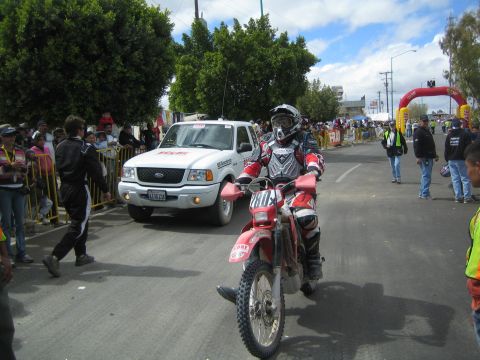 1990 Ford Bronco - racer on a red dirt bike numbered 408 rides down a barricaded street with a white Ford Ranger pickup behind.
