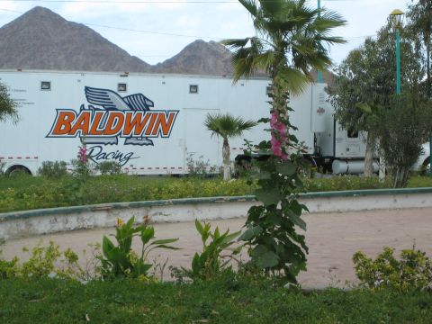 1990 Ford Bronco - white semi-trailer lettered Baldwin Racing is parked beside greenery and a planter with desert mountains behind.