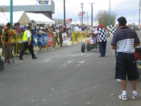 1990 Ford Bronco - racing ATV quad rides down a barricaded street as an official waves a checkered flag and spectators watch.