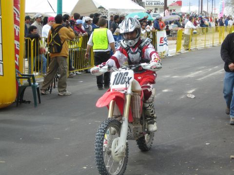1990 Ford Bronco - racer on a red dirt bike numbered 106x rides down a barricaded street past spectators at an off-road event.
