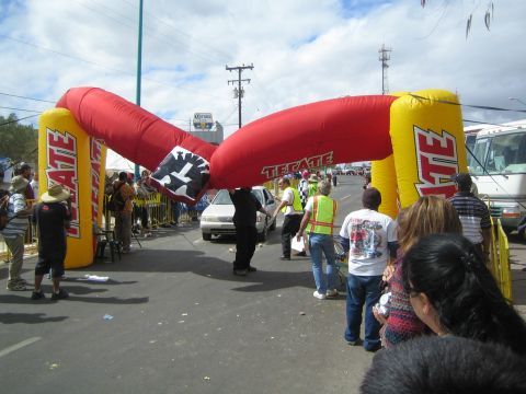 1990 Ford Bronco - red and yellow inflatable Tecate arch spans a street crowded with spectators beside a parked RV.