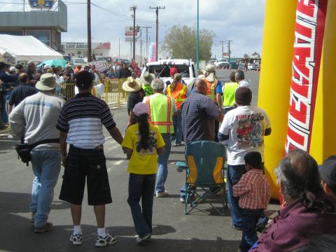 1990 Ford Bronco - Spectators in hats watch from behind a barricaded race street near a yellow Tecate banner and parked vans.