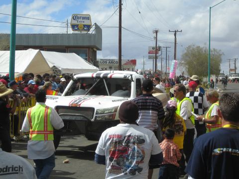1990 Ford Bronco - white off-road race truck is surrounded by crew and spectators on a street beneath a Corona Extra sign.