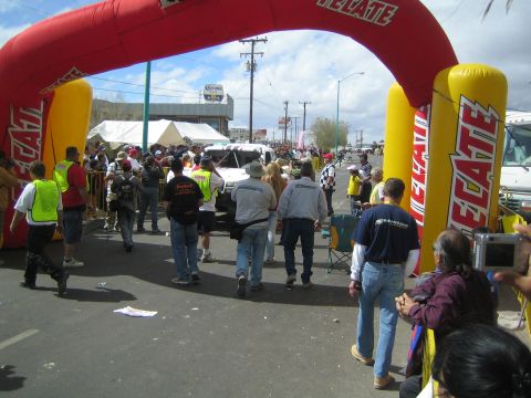 1990 Ford Bronco - Officials and spectators gather under a red and yellow Tecate inflatable arch as a white race truck approaches.