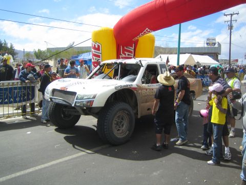 1990 Ford Bronco - white off-road trophy truck on BFGoodrich tires sits under a Tecate arch as crew and fans crowd around.