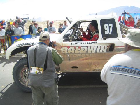 1990 Ford Bronco - photographer and onlookers crowd a mud-spattered white off-road race truck numbered 97 lettered Baldwin Motorsports at a desert event.