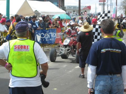 1990 Ford Bronco - red racing quad rolls down a fenced town street as a checkered flag waves and a SCORE official watches the crowd.