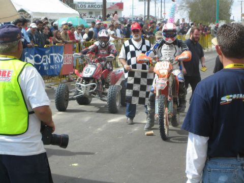 1990 Ford Bronco - numbered racing quad and an orange dirt bike pause at a finish where a man holds a large checkered flag amid spectators.