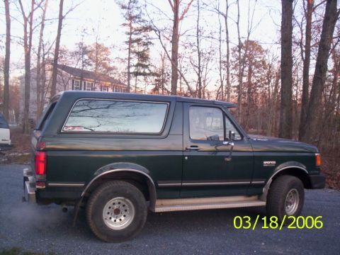 1988 Ford Bronco - This dark green Ford Bronco is viewed from the side, with a house and trees in the background.
