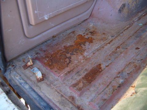 1988 Ford Bronco - The interior floor of a vehicle shows significant rust and wear, with a brown plastic panel visible above.