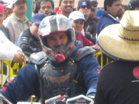 1990 Ford Bronco - grimy racer in a battered full-face helmet and chest protector sits among a packed crowd at a desert race finish.