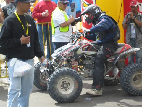 1990 Ford Bronco - helmeted racer in muddy gear leans over a sponsor-covered racing quad as a bystander and officials look on.