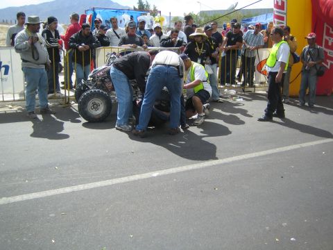1990 Ford Bronco - cluster of officials and crew crouch around a racing quad on a sunlit paved street near barricades and a red inflatable arch.