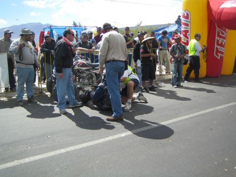 1990 Ford Bronco - Officials and bystanders surround a racing quad on a road beside a red inflatable arch with mountains in the background.