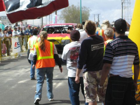 1990 Ford Bronco - crowd in safety vests watches a red and yellow flame-painted off-road race truck approach as a US flag waves overhead.