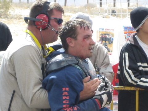1990 Ford Bronco - crew member wearing headphones supports a dazed racer with a cut face and chest armor, jersey lettered Hancock, near a race tent.