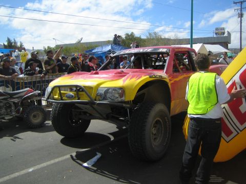 1990 Ford Bronco - red and yellow flame-painted off-road race truck with a roll cage idles on a street as a yellow-vested worker stands beside it.