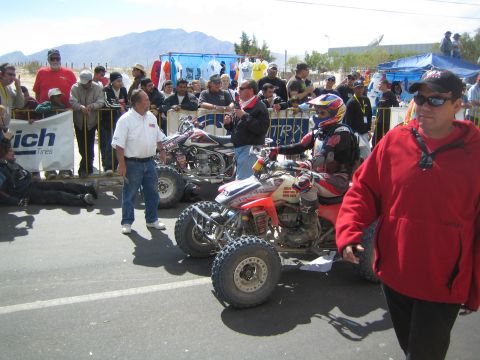 1990 Ford Bronco - Racing quads and riders gather on a barricaded street lined with spectators and laundry, mountains rising behind.