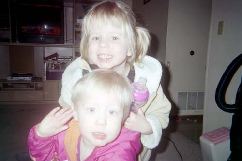 Two young children, one in a pink jacket, the other in a cream-colored coat, indoors