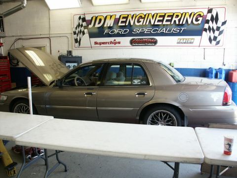 2001 Mercury Grand Marquis - A tan 2001 Mercury Grand Marquis sedan is parked inside a garage with its hood open, under a sign for JDM Engineering.
