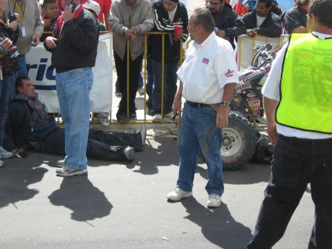1990 Ford Bronco - older man in a sponsor-covered shirt walks across a road past a racing quad and a racer lying near yellow barricades.