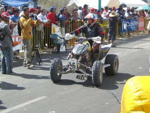 1990 Ford Bronco - helmeted rider pilots racing quad number 56 down a street flanked by spectators behind a yellow barricade fence.