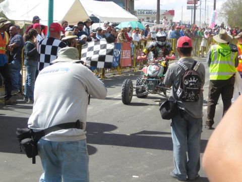 1990 Ford Bronco - Photographers and a checkered flag greet a helmeted rider on a racing quad approaching the finish on a town street.