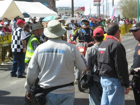1990 Ford Bronco - dense crowd including a man in a Quiksilver shirt surrounds a helmeted dirt bike rider at a barricaded town finish.