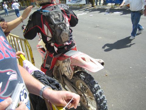 1990 Ford Bronco - muddy rear of a dirt bike and its armored rider sit beside a barricade as spectators reach over the rail.
