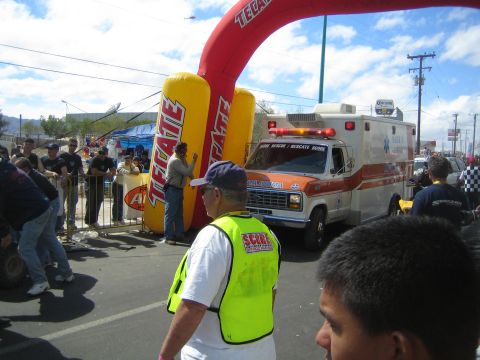 1990 Ford Bronco - orange and white ambulance parks beside a Tecate inflatable arch as a SCORE official in a vest stands at a desert race.