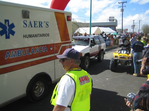1990 Ford Bronco - white Saer's Ambulancia and an SUV sit beside a racing quad on a street as a SCORE official watches the finish crowd.