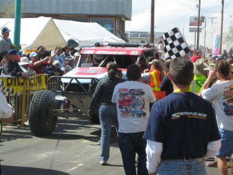 1990 Ford Bronco - red off-road race truck numbered 102 inches through a town crowd as a checkered flag waves at the finish.