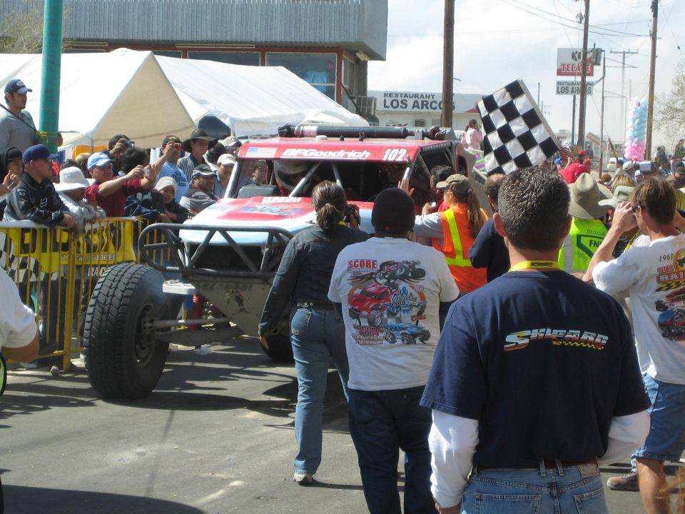 Red off-road race truck number 102 surrounded by a crowd and a checkered flag on a town street