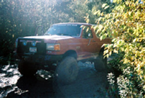 2002 Oldsmobile Aurora - An orange pickup truck navigates a wooded trail with large tires.