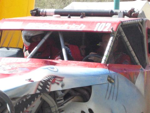 1990 Ford Bronco - shark-mouth-painted red race truck numbered 102 sits near tents with two helmeted occupants visible inside the cage.