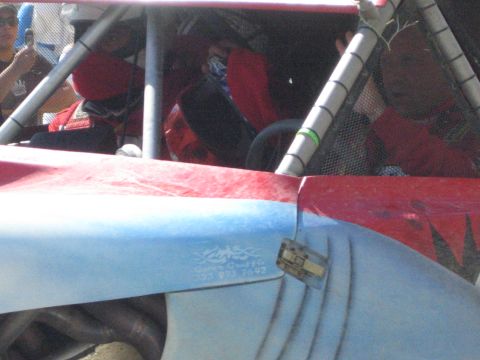 1990 Ford Bronco - View into the cockpit of a red off-road buggy showing seated occupants, steering wheel and roll cage, with a bystander filming on a phone.