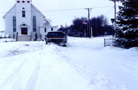 1984 Ford Bronco - A red SUV is stuck in deep snow in front of a white church on a cloudy day.
