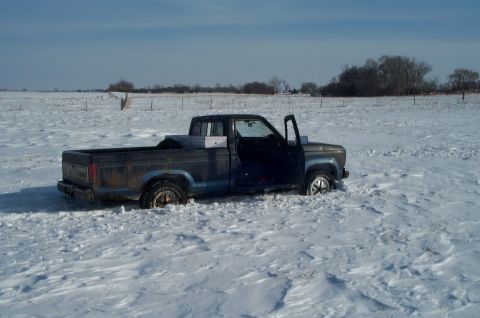 1984 Ford Bronco - Pickup truck stuck in a snowy field with the driver's door ajar.