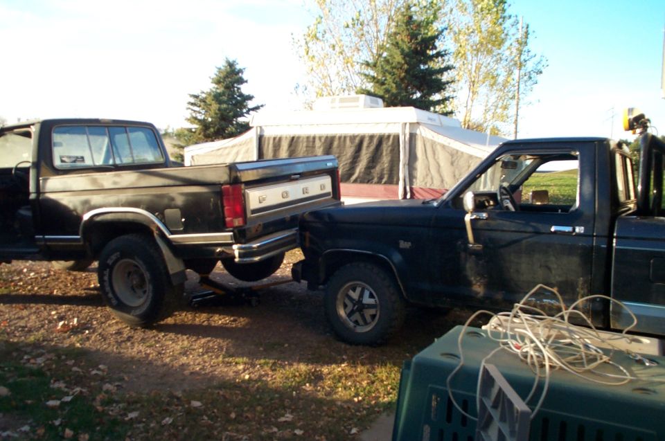 Two older black Ford pickup trucks parked outdoors near a tent and trees at dusk