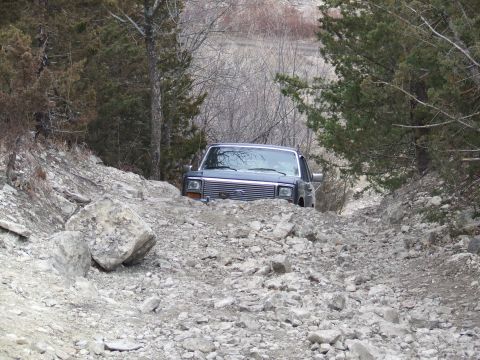 1986 Ford Bronco - Ford Bronco on a challenging off-road trail, surrounded by rocks and trees.