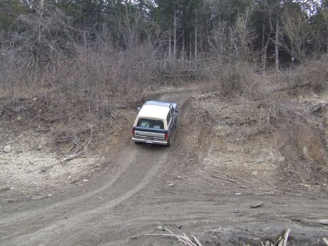1986 Ford Bronco - A two-tone Ford Bronco navigates an off-road dirt path surrounded by bare trees.