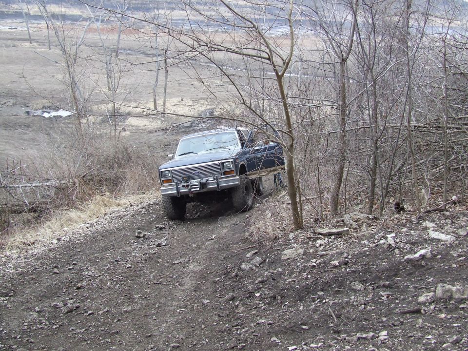 Dark blue Ford Bronco driving up a dirt hill with trees on the right