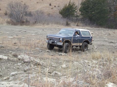 1986 Ford Bronco - A dark blue Ford Bronco with a white top navigates a rocky, off-road path.