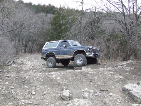 1986 Ford Bronco - Ford Bronco navigates a challenging off-road course with dense trees and rocky terrain.