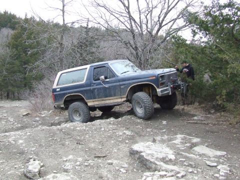 1986 Ford Bronco - A lifted Ford Bronco tackles a challenging off-road course with a person standing nearby.