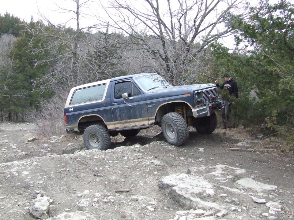 Dark blue Ford Bronco with a white top, lifted suspension, and off-road tires, navigating rocky terrain