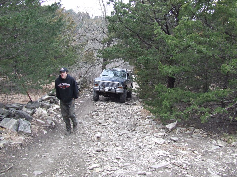 Man in black hoodie and camouflage pants stands on a rocky trail next to a 1986 Ford Bronco